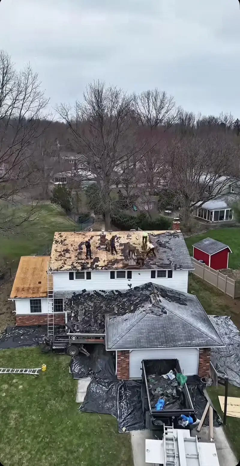 Aerial view of a crew tearing off old shingles with a dumpster and tarps protecting the lawn